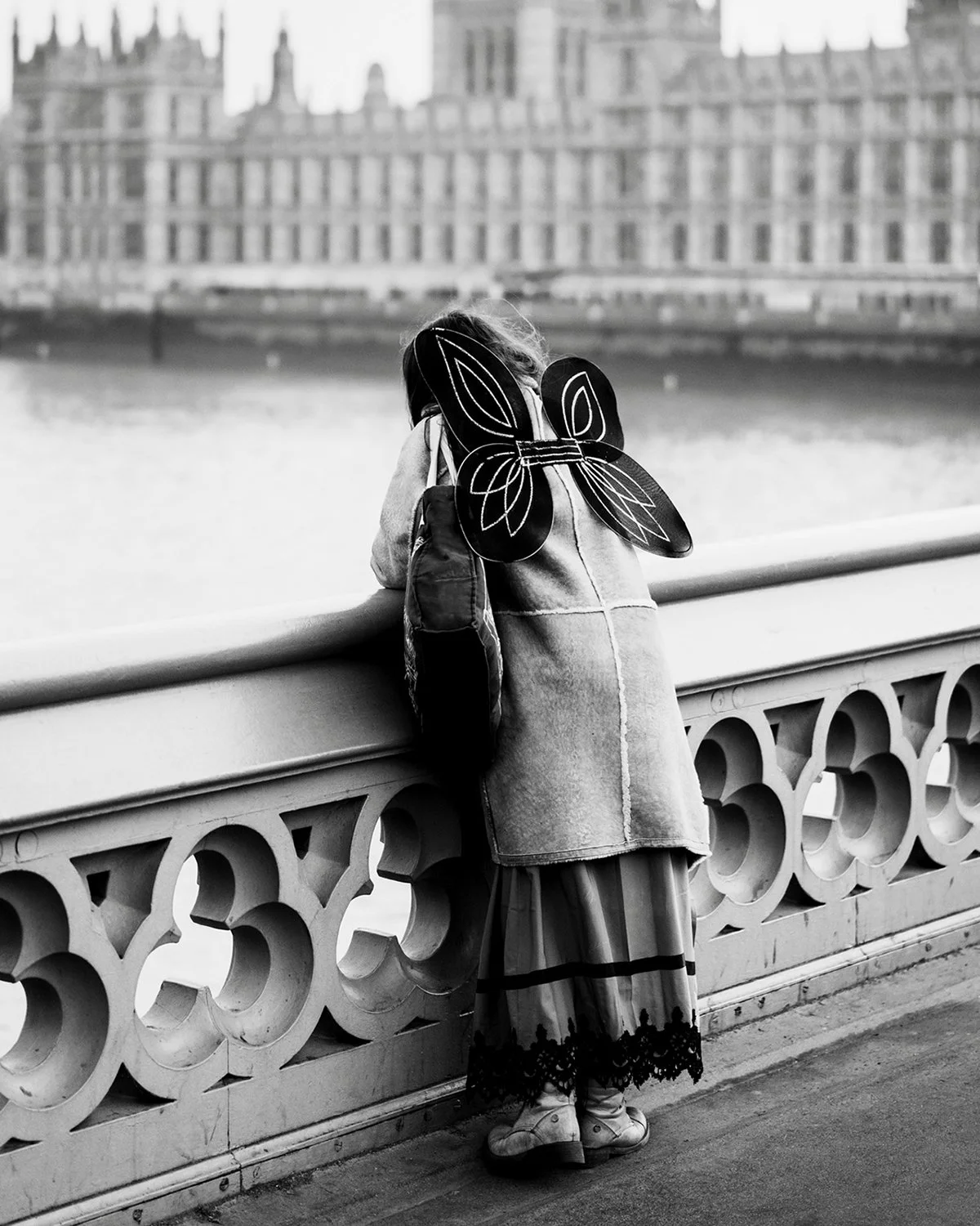A person with butterfly wings costume leaning on a bridge railing along a river, with historic buildings in the background.