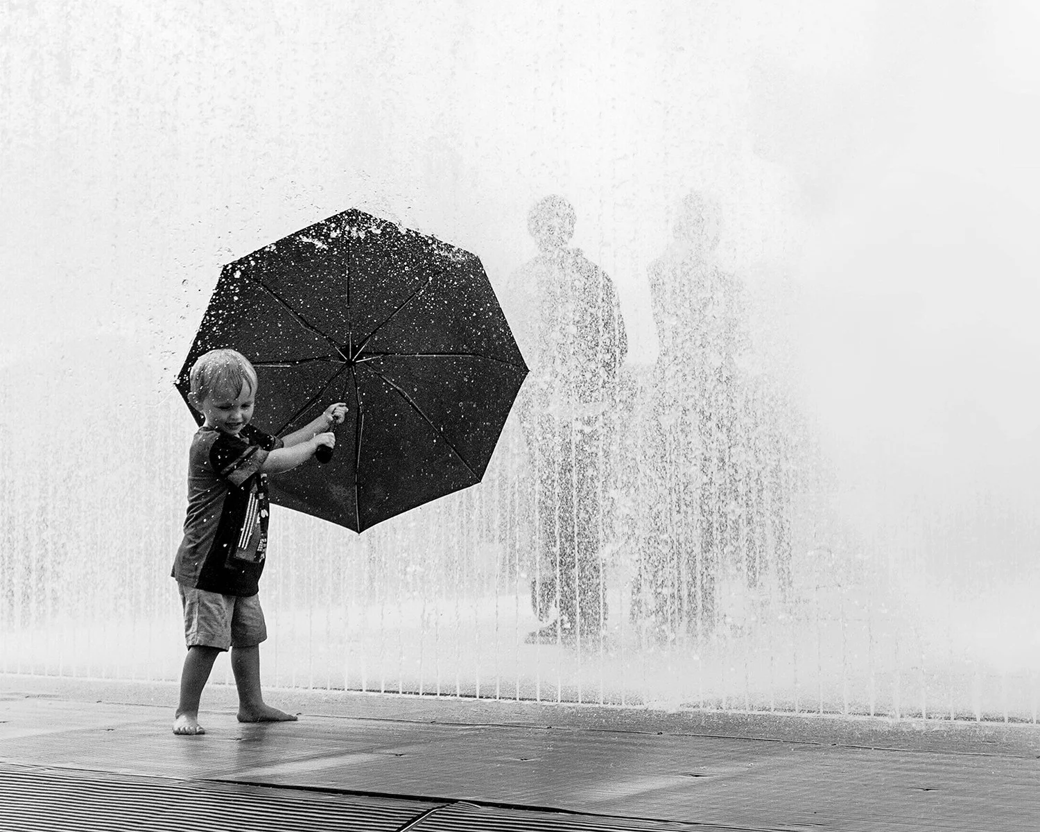 A young boy holding an umbrella in front of a fountain, with two blurred adults in the background.