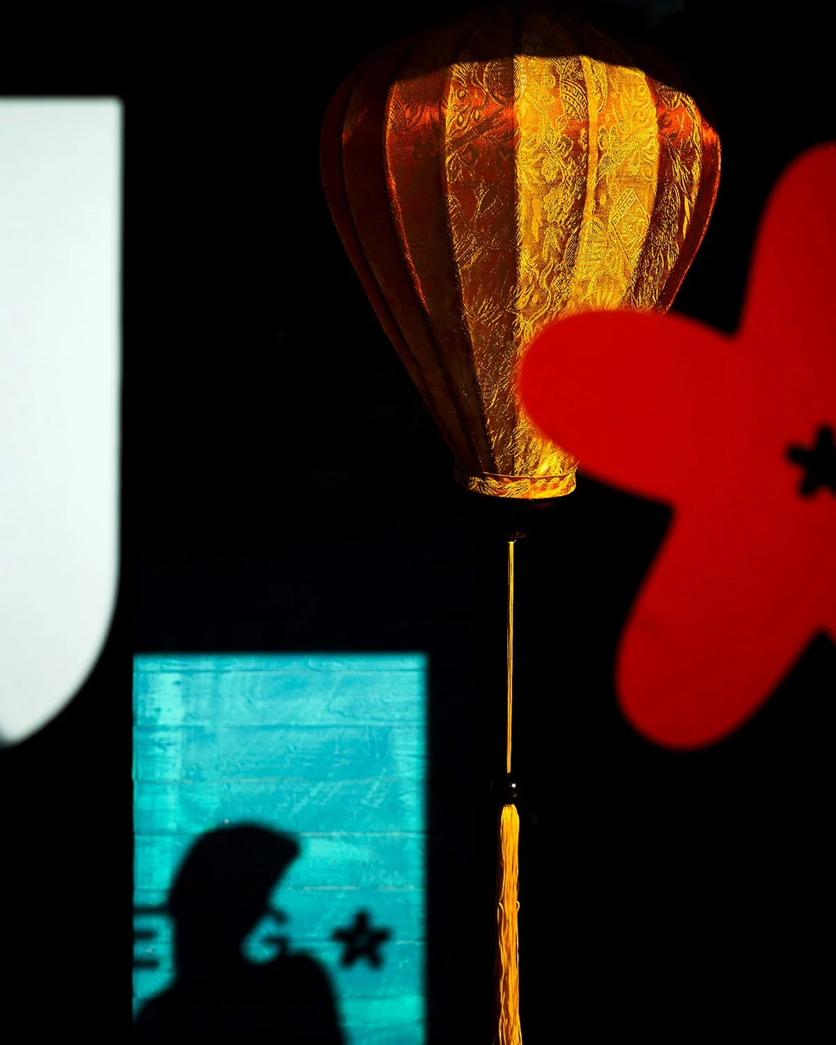 Illuminated paper lantern with gold and red colors, partially obscured by red flower-shaped decoration, against a dark background.