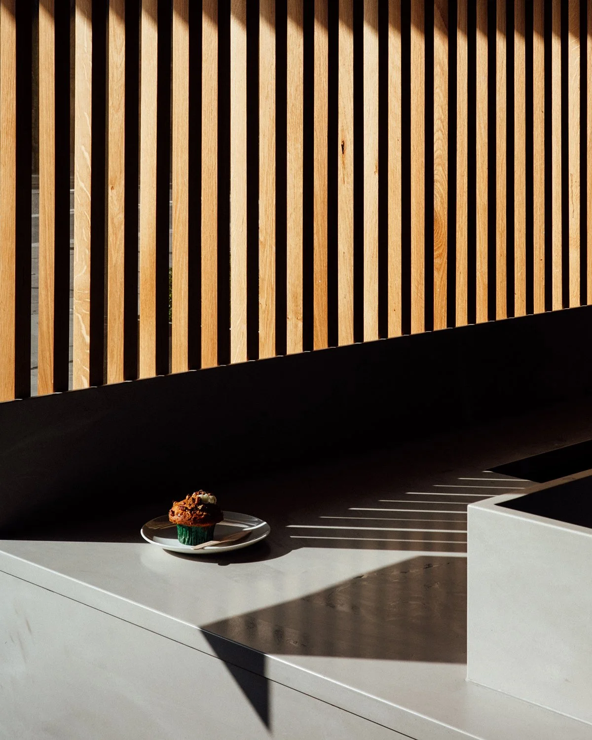 A single cupcake with frosting on a white plate on a light-colored surface, with shadows and sunlight creating striped patterns. A wooden slatted wall is in the background.