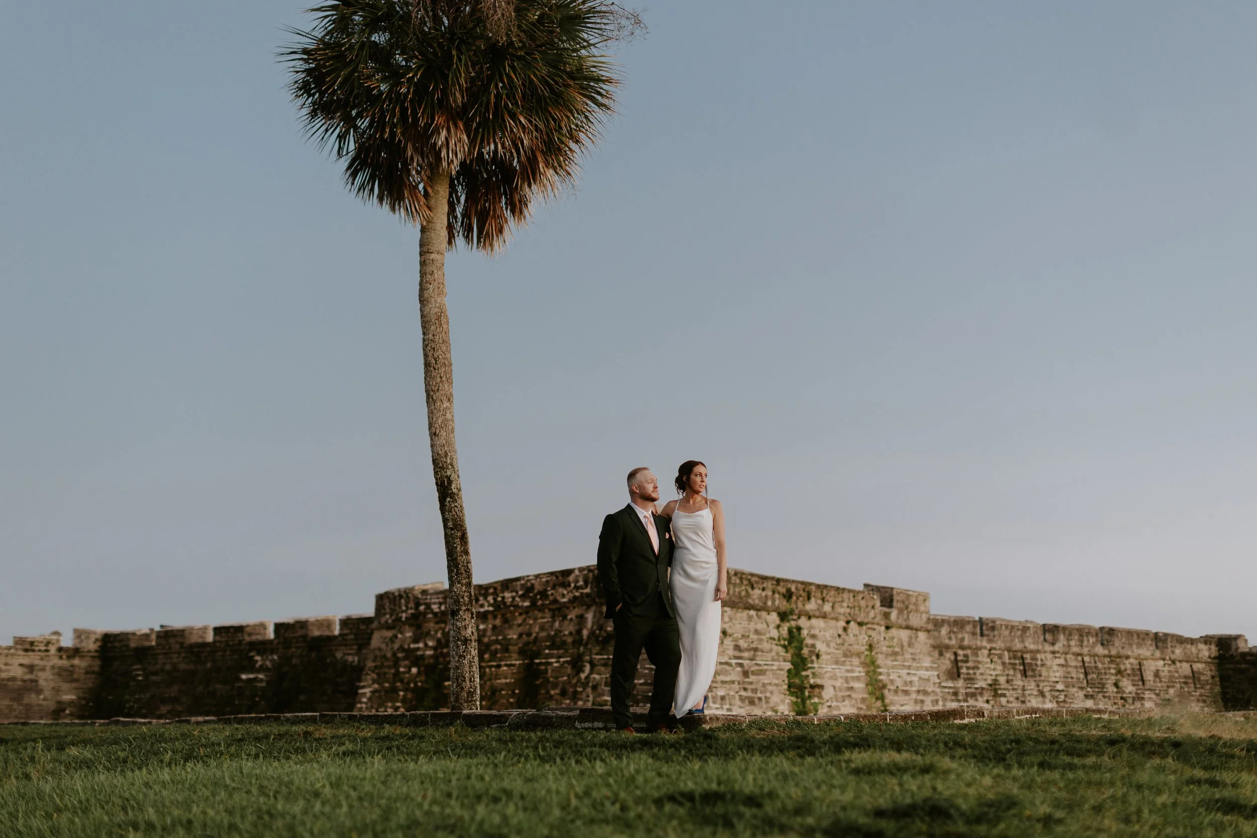 Wedding couple posing near a historic stone fort and palm tree during golden hour in St. Augustine, Florida.