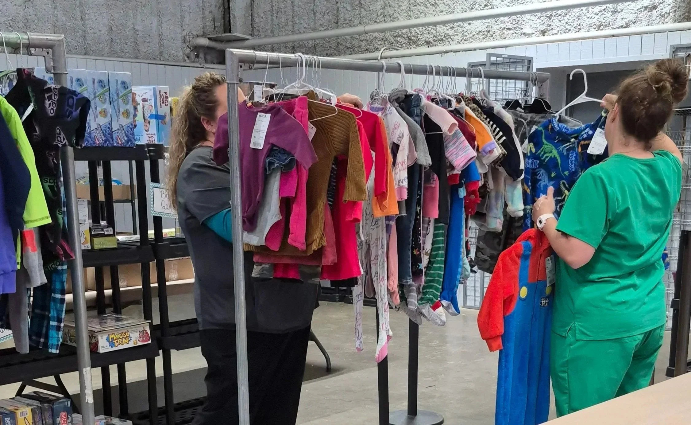 Image shows 2 women looking through a rack of children's clothing and inspecting them for stains, tears, or missing pieces