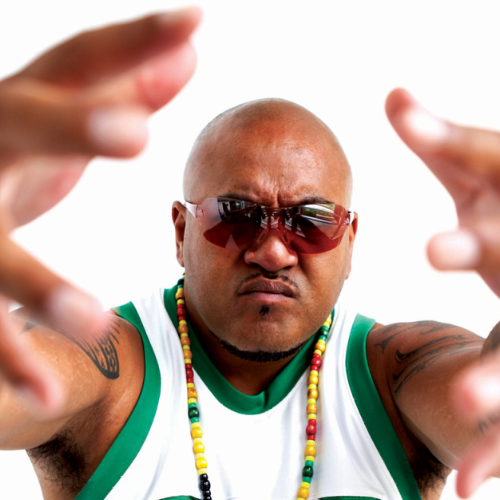 A bald man wearing sunglasses, a white tank top with green trim, and colorful beaded necklaces, looking intently at the camera with his arms extended forward.