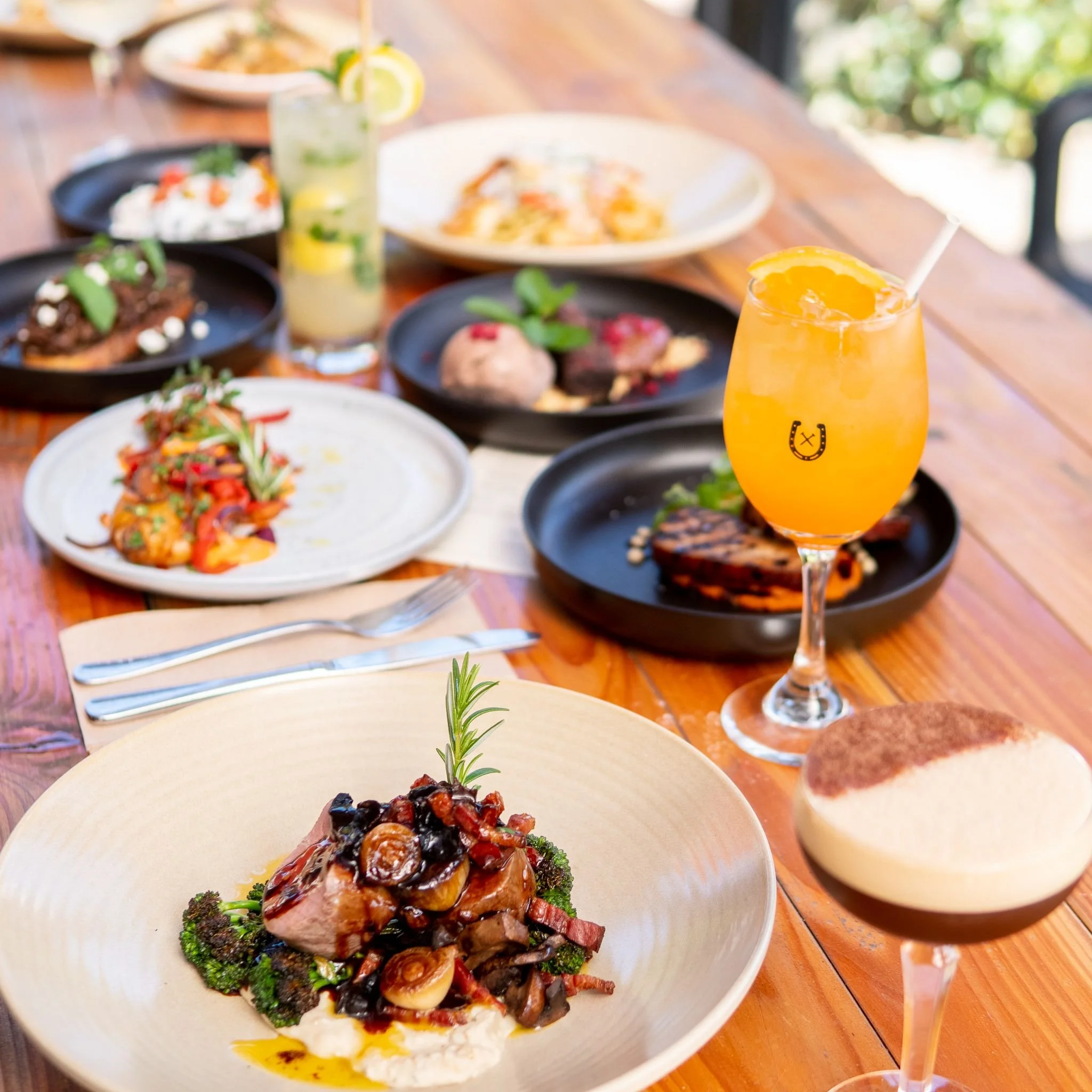 A wooden table set with various gourmet dishes and drinks, including a plate with meat and vegetables garnished with rosemary, an orange cocktail, and a coffee-flavored beverage in a coupe glass. Several other plates with assorted appetizers and entrees are also visible in the background.