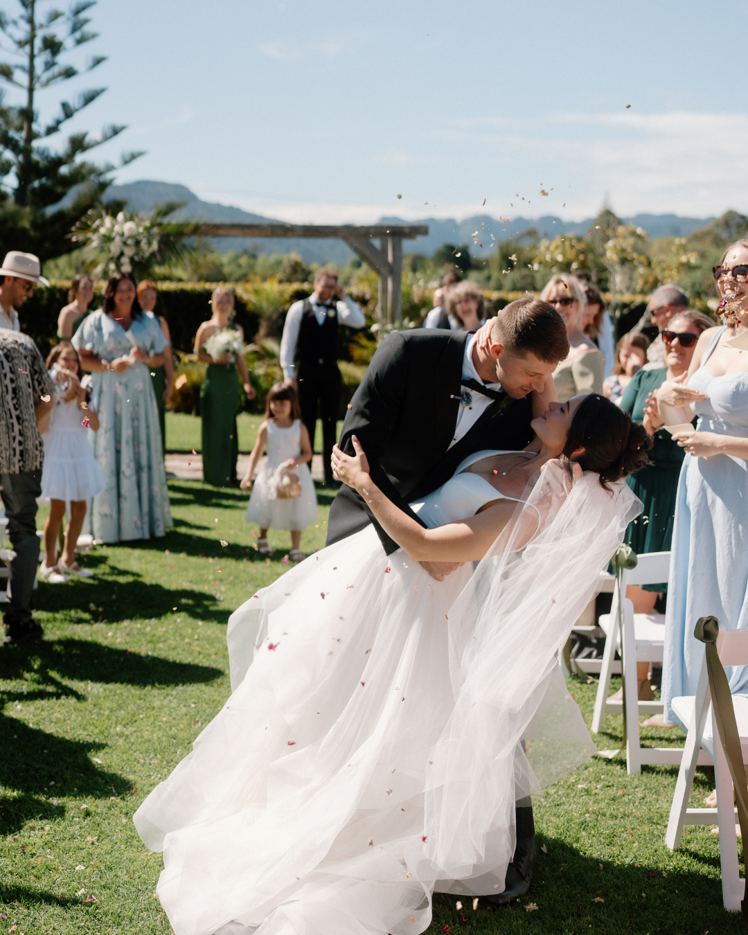 Bride and groom kissing at outdoor wedding ceremony under a decorated arch, while guests watch and clap.