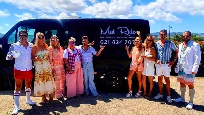 Group of people in colorful outfits posing in front of a black van with "Mai Ride" branding, outdoors on a sunny day.