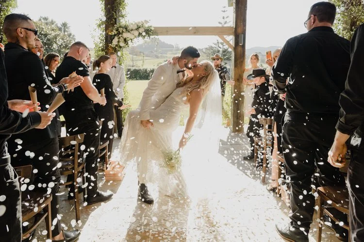 Bride and groom kissing at the end of the wedding aisle, surrounded by guests throwing confetti, with a scenic outdoor background.