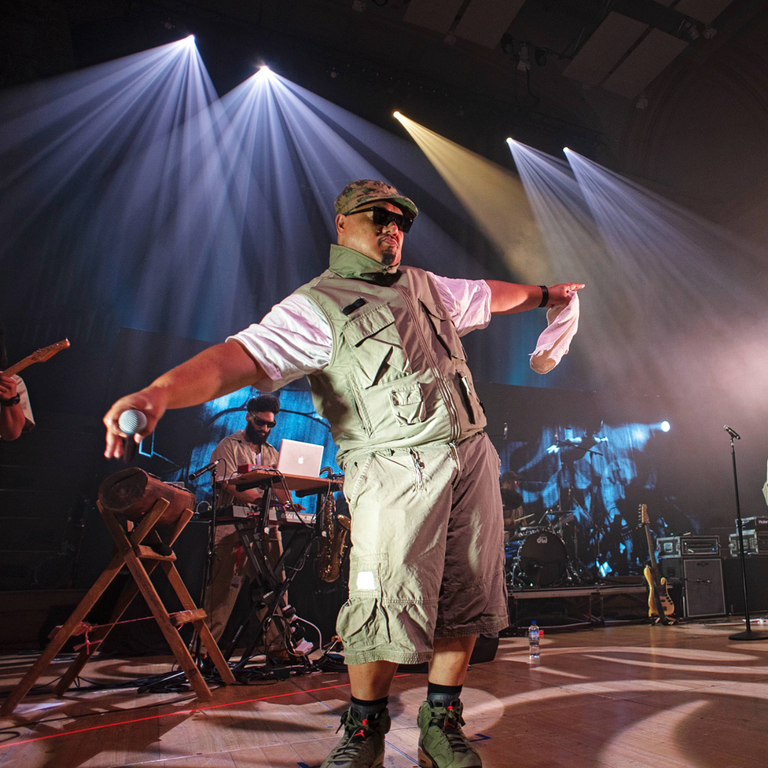 Performer dancing on stage with musicians and equipment in the background, colorful stage lights overhead.