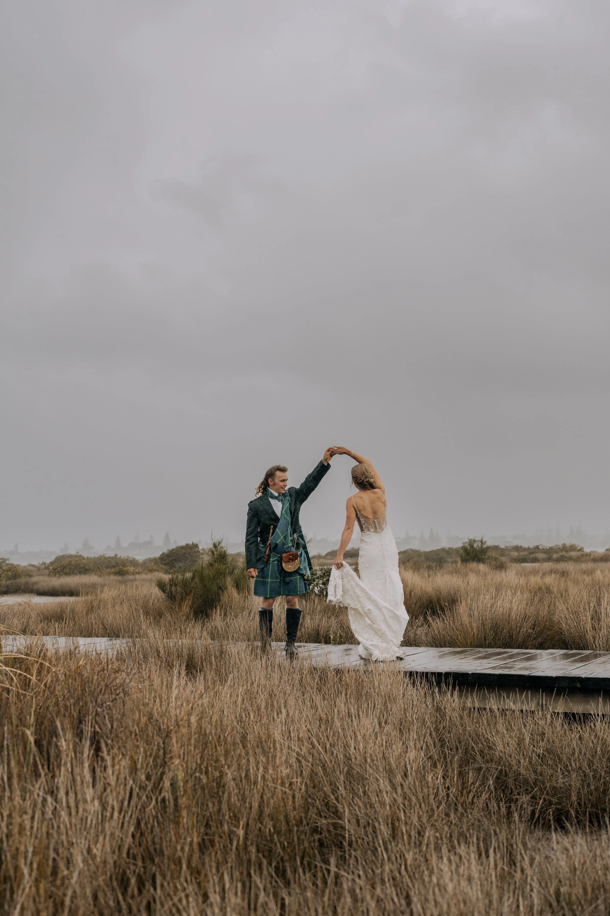 A wedding ceremony with a bride in a white dress holding hands with the groom in a blue suit, in front of a rustic stone fireplace with floral decorations. Bridesmaids and groomsmen are in attendance.