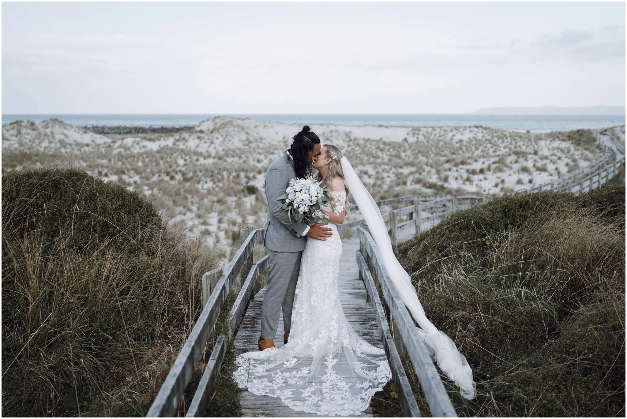 Bride and groom kissing on a beach boardwalk, with sand dunes and ocean in the background.