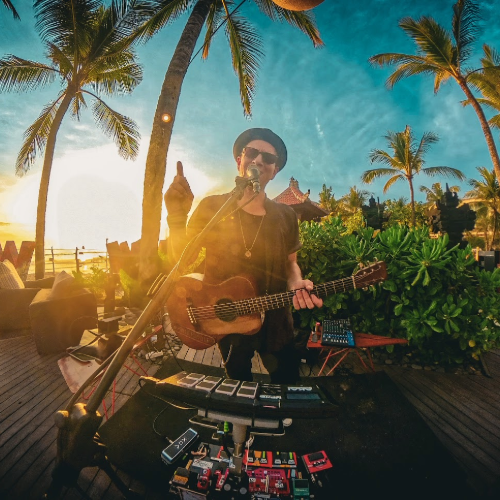 Musician performing outdoors on a tropical beach at sunset, playing guitar and singing into a microphone, surrounded by palm trees and lush greenery.