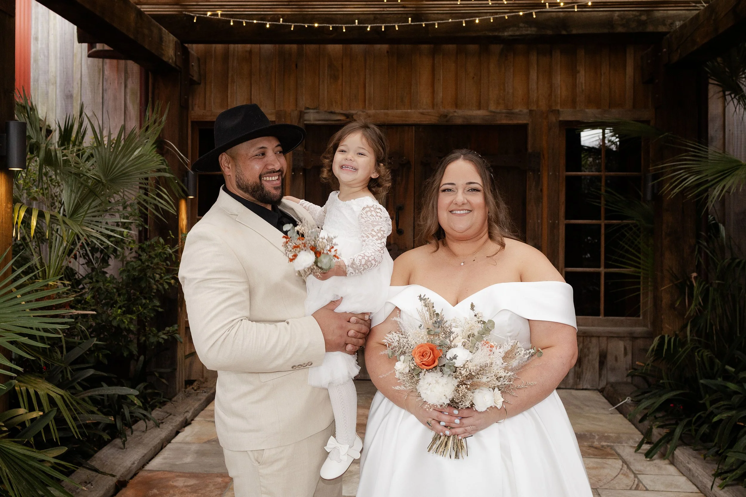 A wedding ceremony with a bride in a white dress holding hands with the groom in a blue suit, in front of a rustic stone fireplace with floral decorations. Bridesmaids and groomsmen are in attendance.