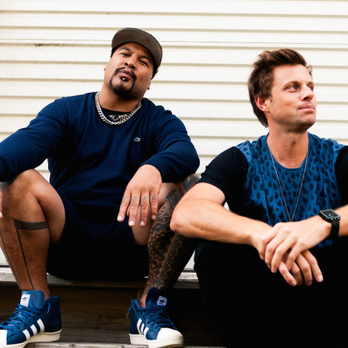 Two men sitting outdoors in front of a white vinyl siding wall, dressed casually and looking away, one with a serious expression and the other with a relaxed look.