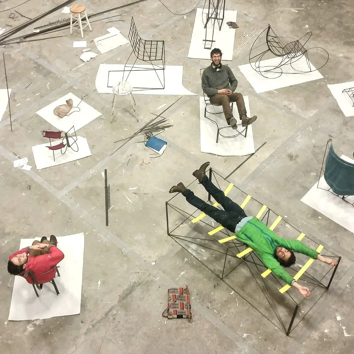 Aerial view of a spacious studio with various wireframe chairs and stools on white papers, three people sitting and one lying down