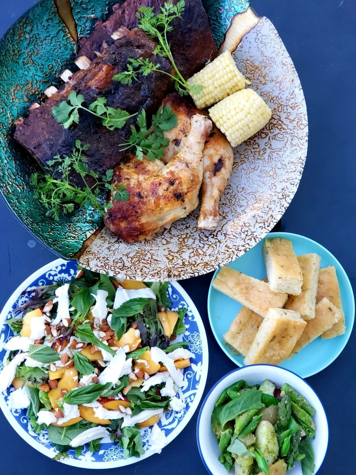Overhead flatlay of four colorful and delicious catering dishes: a fresh salad, focaccia, smoked ribs, and chicken legs with corn.