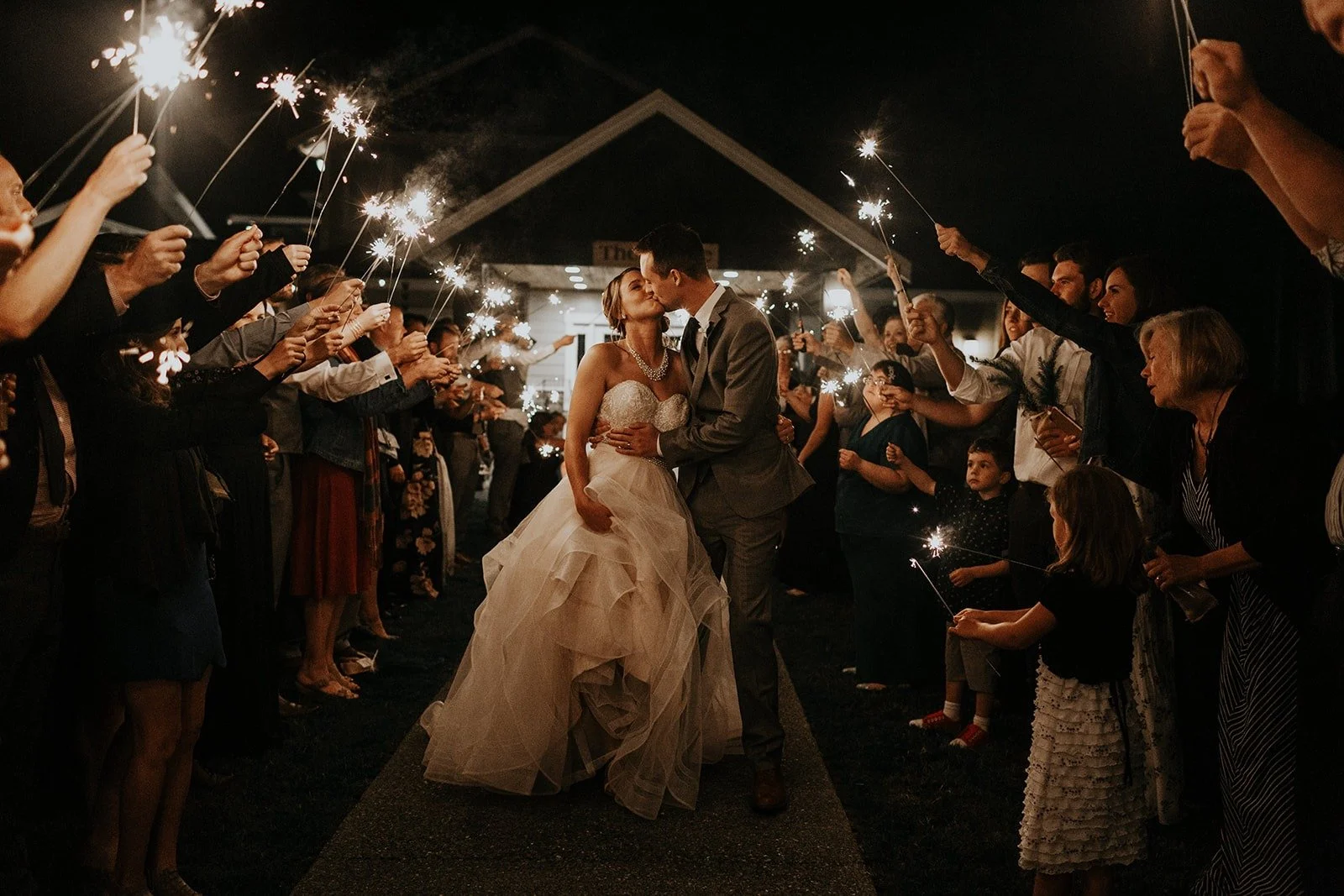 Bride and groom kissing at dusk as wedding guests hold sparklers in an aisle, creating a tunnel of light for the couple's grand exit.