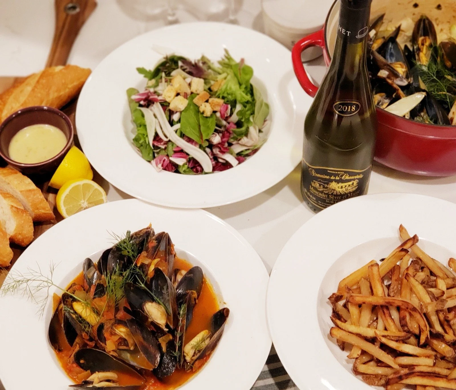 An elegant and refined holiday celebratory dinner setting featuring gourmet dishes like vongole in sauce, a colorful salad, a potato side, and a 2018 sparkling wine.
