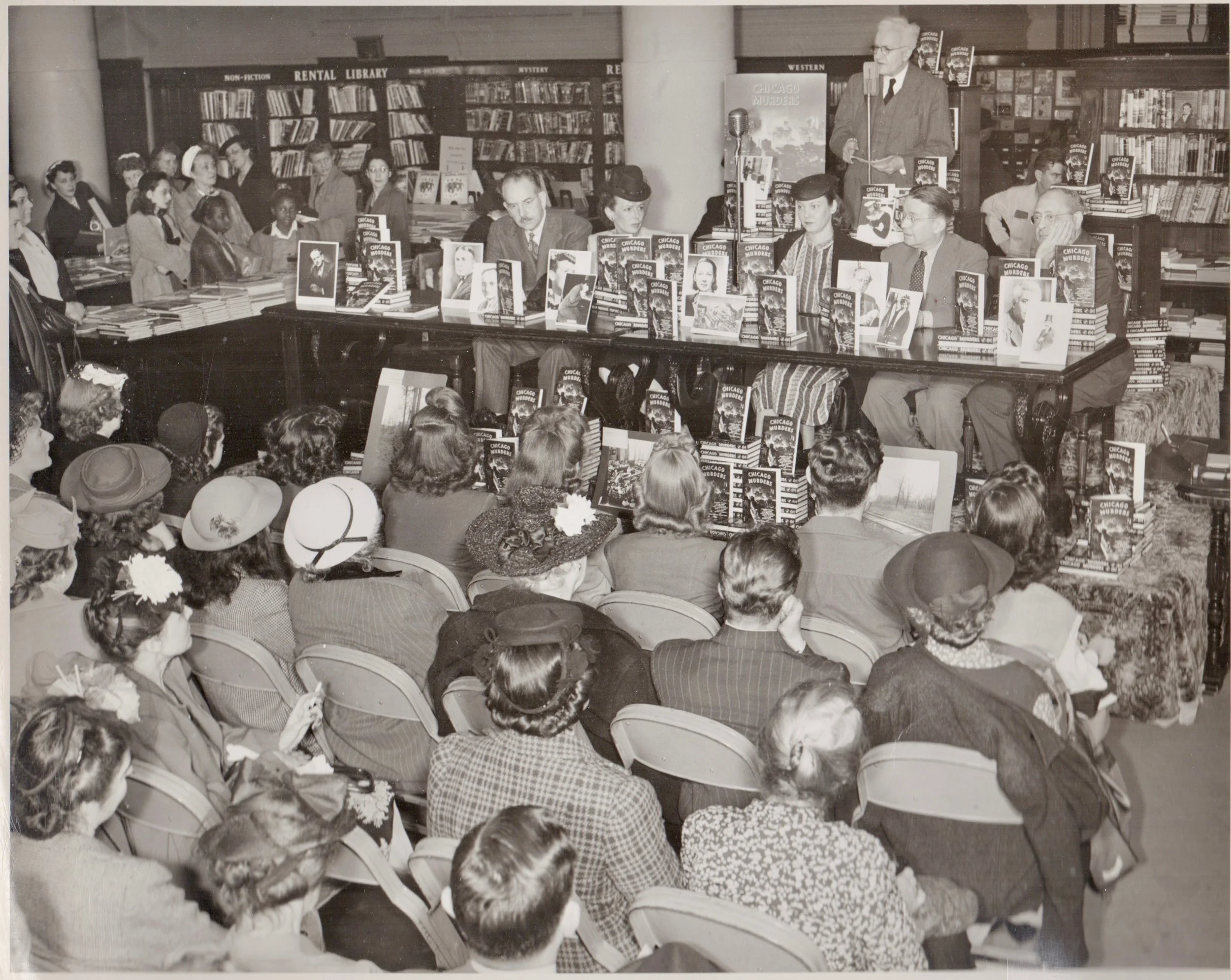 An autograph party featuring the writers of the anthology Chicago Murder . The event took place in June 1945 at the book department of Marshall Field & Co. in downtown Chicago. Look at that packed house! Every seat is full and there is standing room only. They're going to need all those stacks of Chicago Murder !