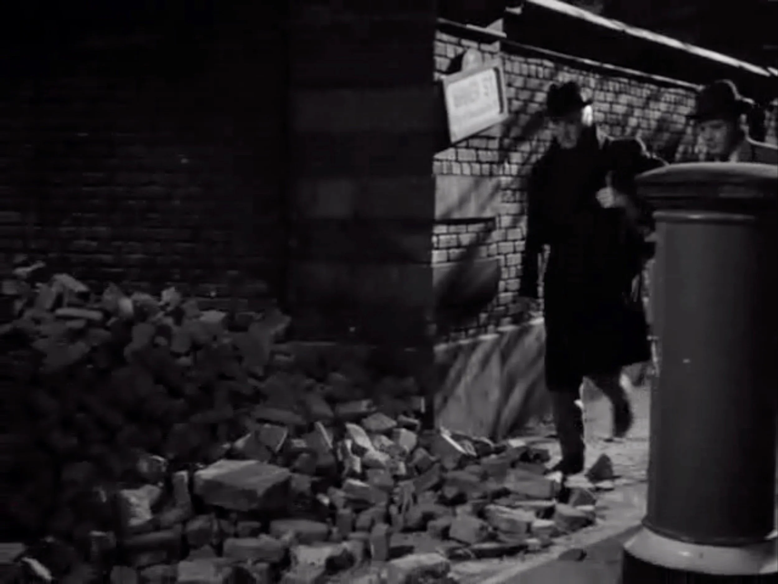 ‘That fabled street.’ In this still image from “Sherlock Holmes and the Secret Weapon,” the battered Baker Street sign is on the wall just to Rathbone’s left. In this scene, Holmes is escorting Dr. Franz Tobel played by William Post Jr., and his bombsight safely to London. Just after this image, Tobel will slip on some bricks, and Holmes will say that Tobel will need to get used to London’s blackouts.