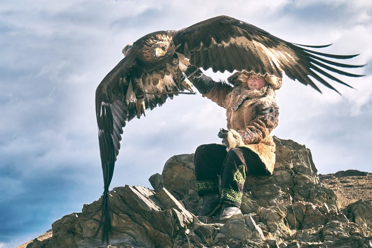 Person in traditional attire holding a large golden eagle on rocky terrain with cloudy sky. Film Mongolia. Filming in Mongolia. Producer in Mongolia. Film Production Service in Mongolia. Fixer Service Mongolia.