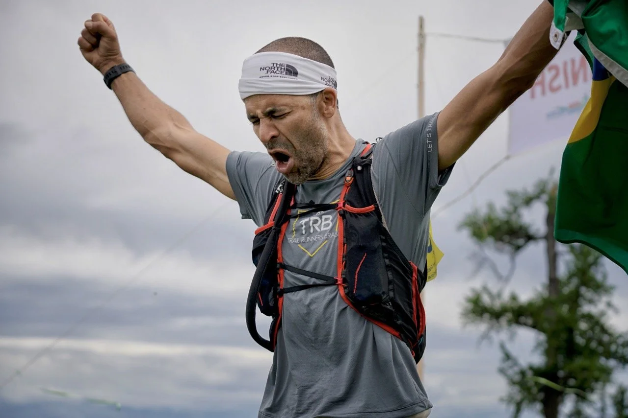 A male runner celebrates with arms raised after finishing a trail race, wearing a headband and hydration vest. A partial view of the Brazilian flag is visible.
