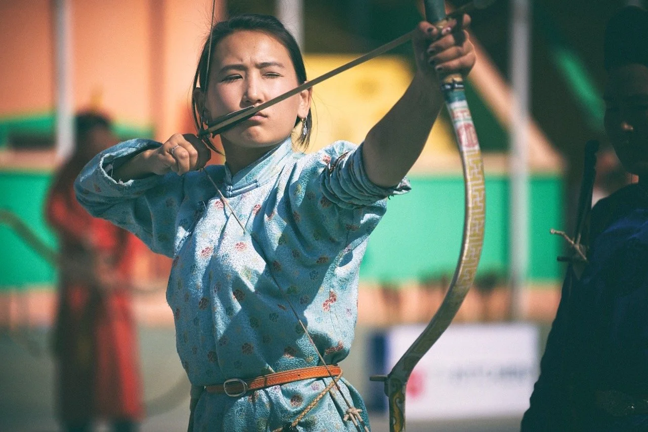 Person in traditional attire aiming a bow and arrow outdoors.Film Mongolia. Filming in Mongolia. Producer in Mongolia. Film Production Service in Mongolia. Fixer Service Mongolia.