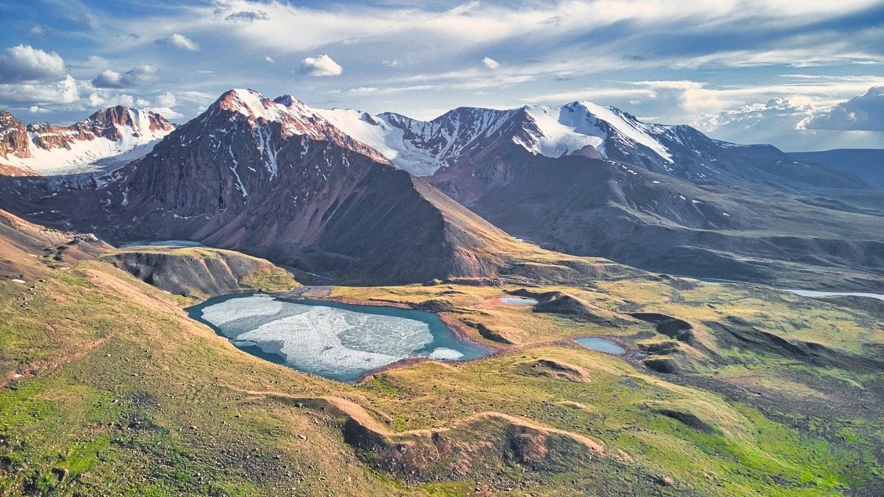 Film Mongolia. Filming in Mongolia. Producer in Mongolia. Film Production Service in Mongolia. Scenic mountain landscape with snow-capped peaks, rocky terrain, and a partially frozen lake in the foreground under a blue sky with clouds. Uvs Province
