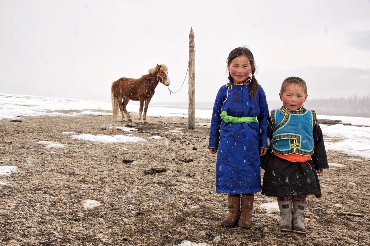 Two children in traditional clothing standing on a snowy field with a brown horse tied to a wooden post in the background. Film Mongolia. Filming in Mongolia. Producer in Mongolia. Film Production Service in Mongolia. Fixer Service Mongolia.