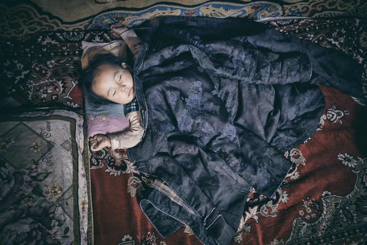 Sleeping child on colorful patterned rugs, wrapped in a dark blanket. Film Mongolia. Filming in Mongolia. Producer in Mongolia. Film Production Service in Mongolia. Fixer Service Mongolia.