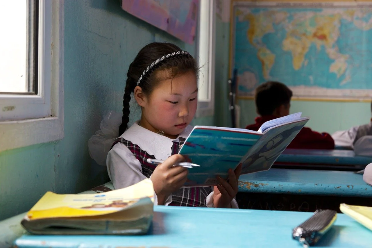 Young girl in school uniform reading a book at a desk in a classroom with a world map on the wall.Film Mongolia. Filming in Mongolia. Producer in Mongolia. Film Production Service in Mongolia. Fixer Service Mongolia.