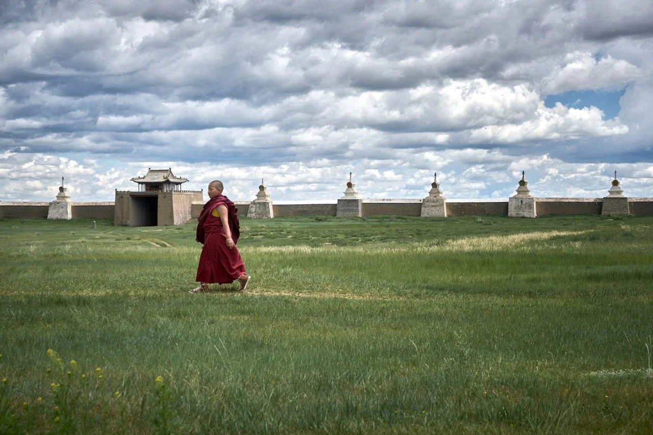 A monk in a red robe walking across a grassy field in front of Erdene Zuu Monastery in Mongolia. Film Mongolia. Filming in Mongolia. Producer in Mongolia. Film Production Service in Mongolia. Fixer Service Mongolia.