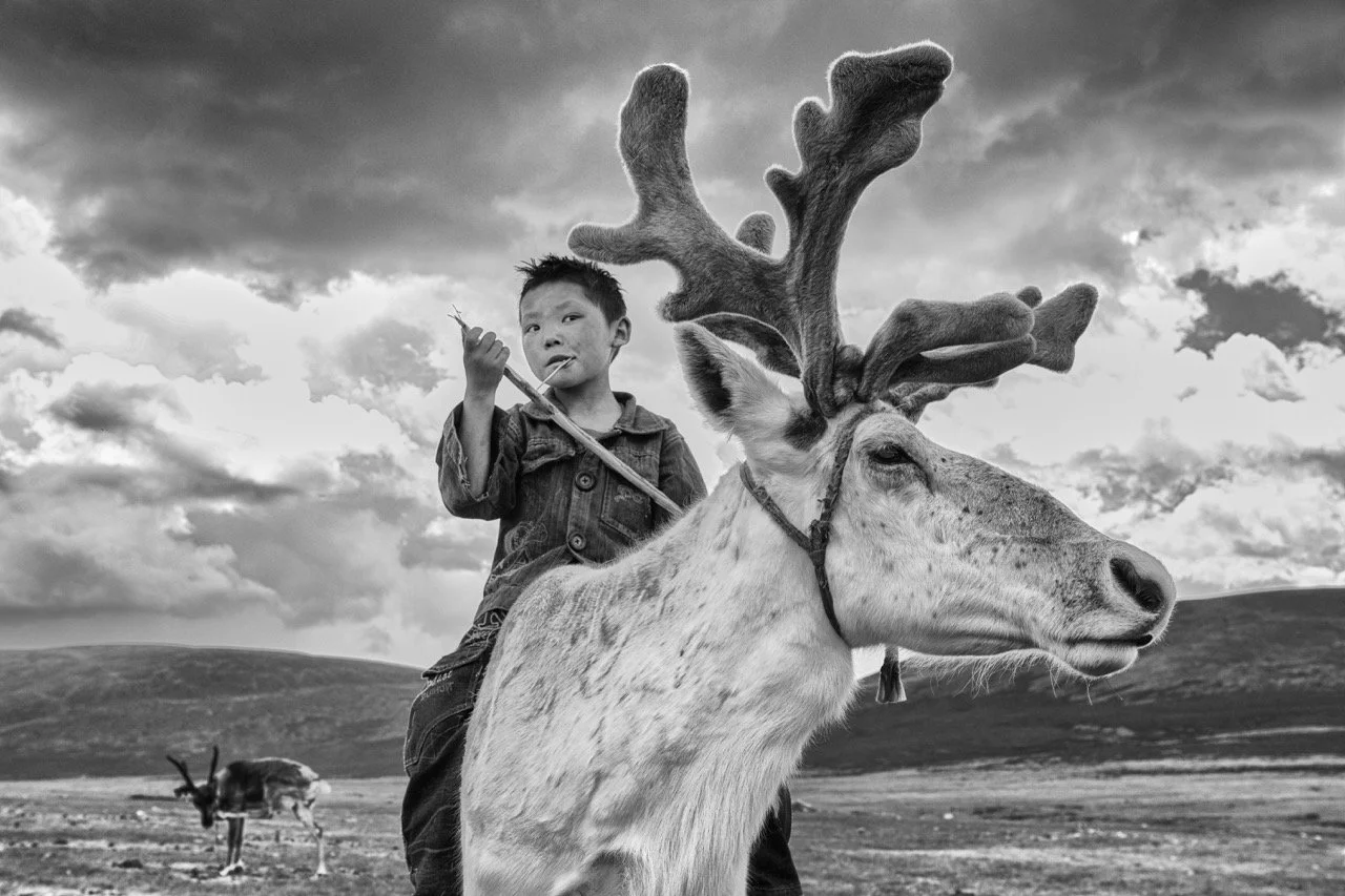 Black and white photo of a young boy sitting on a reindeer in a rugged landscape with overcast skies. Film Mongolia. Filming in Mongolia. Producer in Mongolia. Film Production Service in Mongolia. Fixer Service Mongolia.