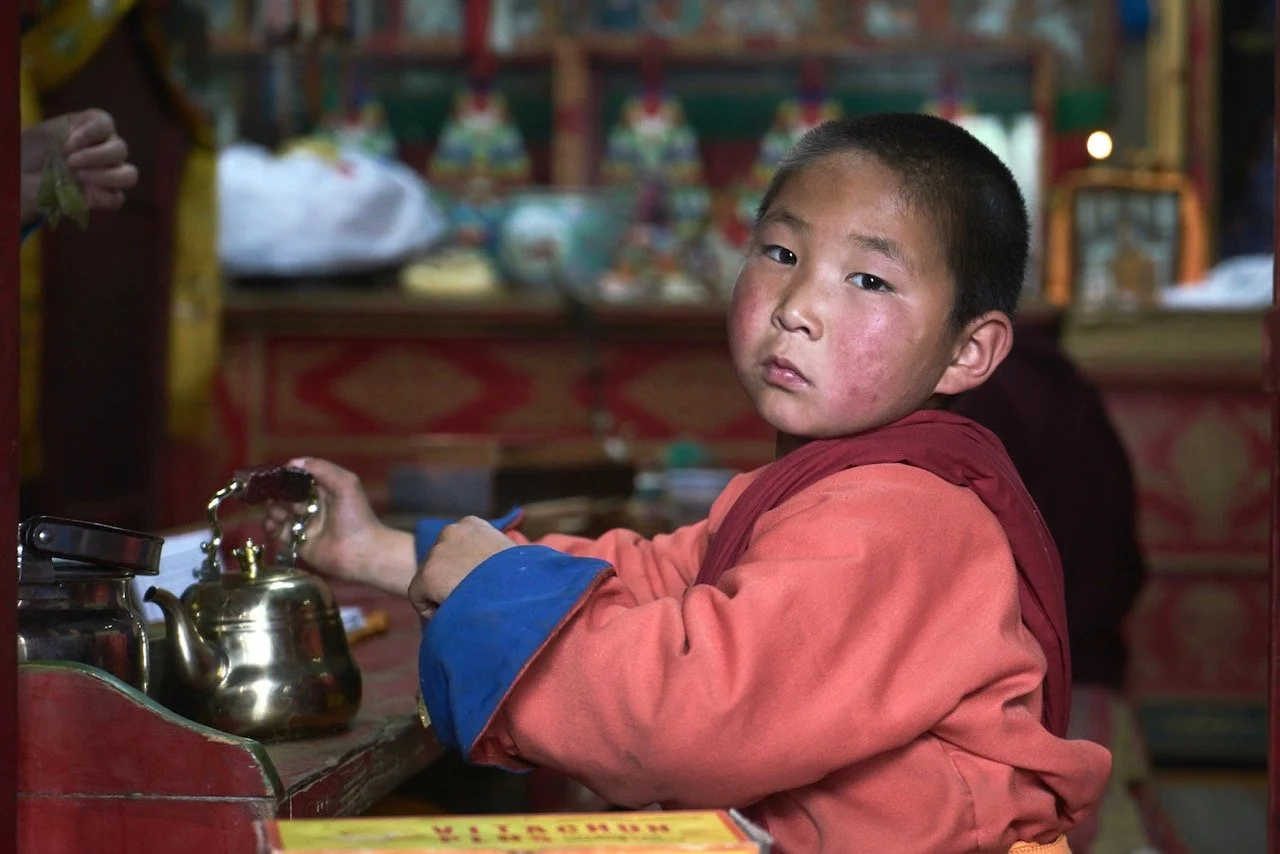 Young monk in traditional robe sitting at a table with a teapot in a temple setting. Kharakhorum, Mongolia. Film Mongolia. Filming in Mongolia. Producer in Mongolia. Film Production Service in Mongolia. Fixer Service Mongolia.