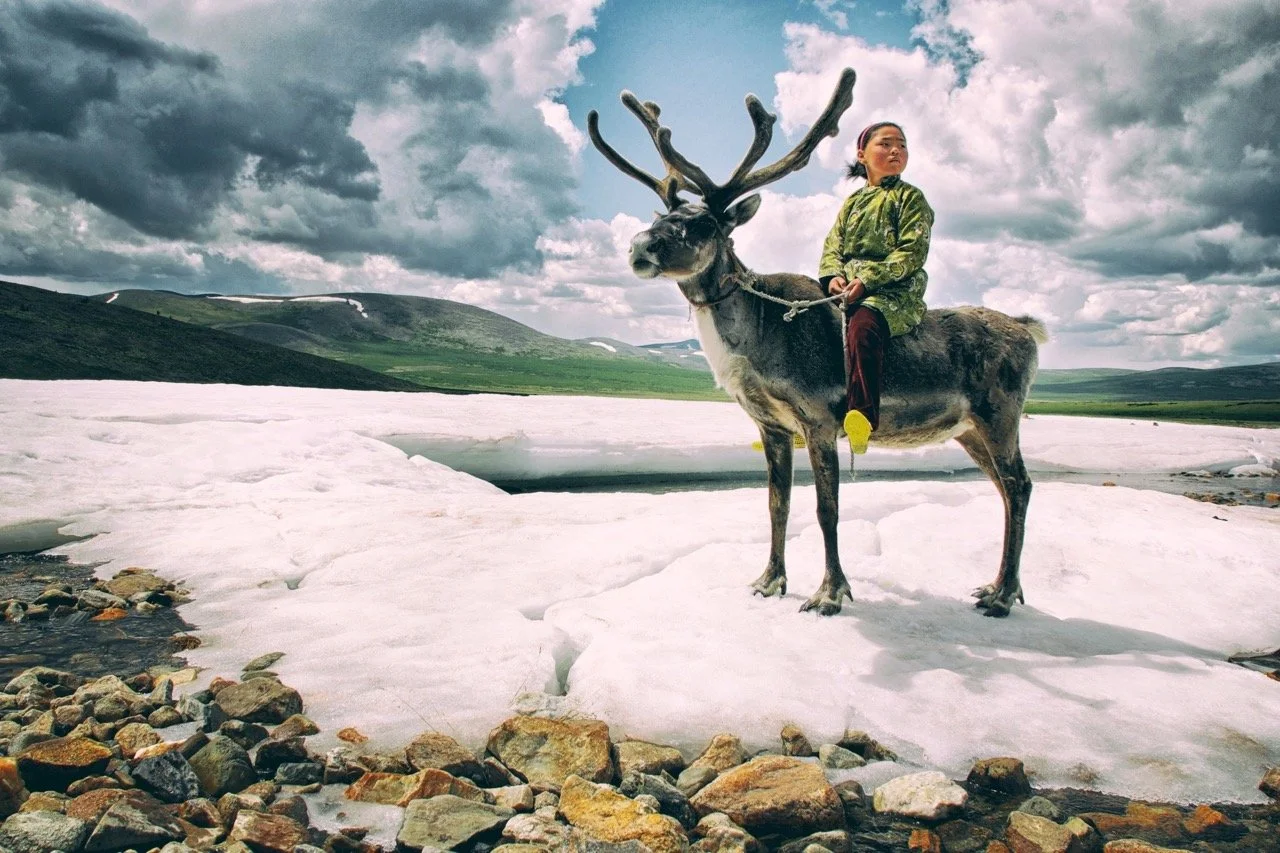 A person in traditional clothing riding a reindeer on a snowy landscape with mountains and a cloudy sky in the background. Film Mongolia. Filming in Mongolia. Producer in Mongolia. Film Production Service in Mongolia. Fixer Service Mongolia.