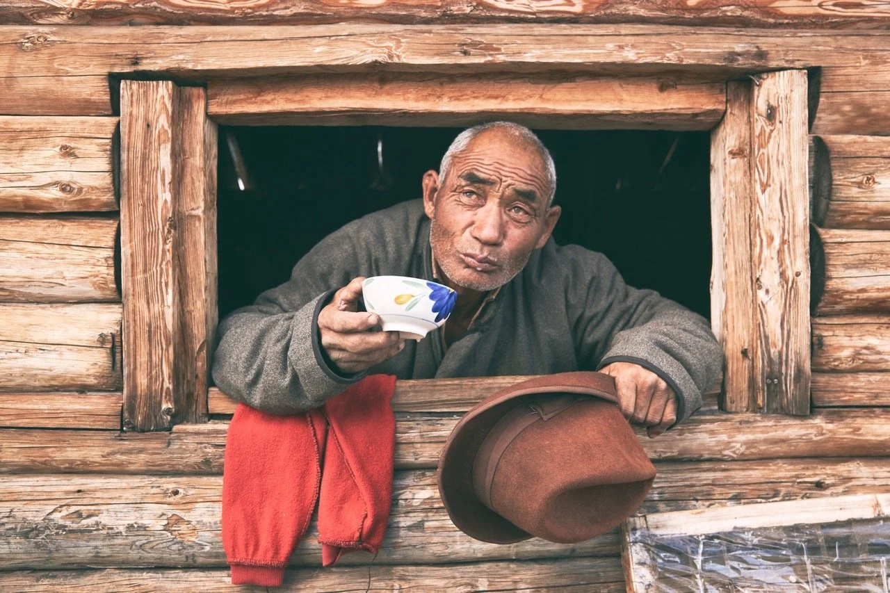 Elderly man holding a cup and hat, leaning out of a wooden window. Film Mongolia. Filming in Mongolia. Producer in Mongolia. Film Production Service in Mongolia. Fixer Service Mongolia.