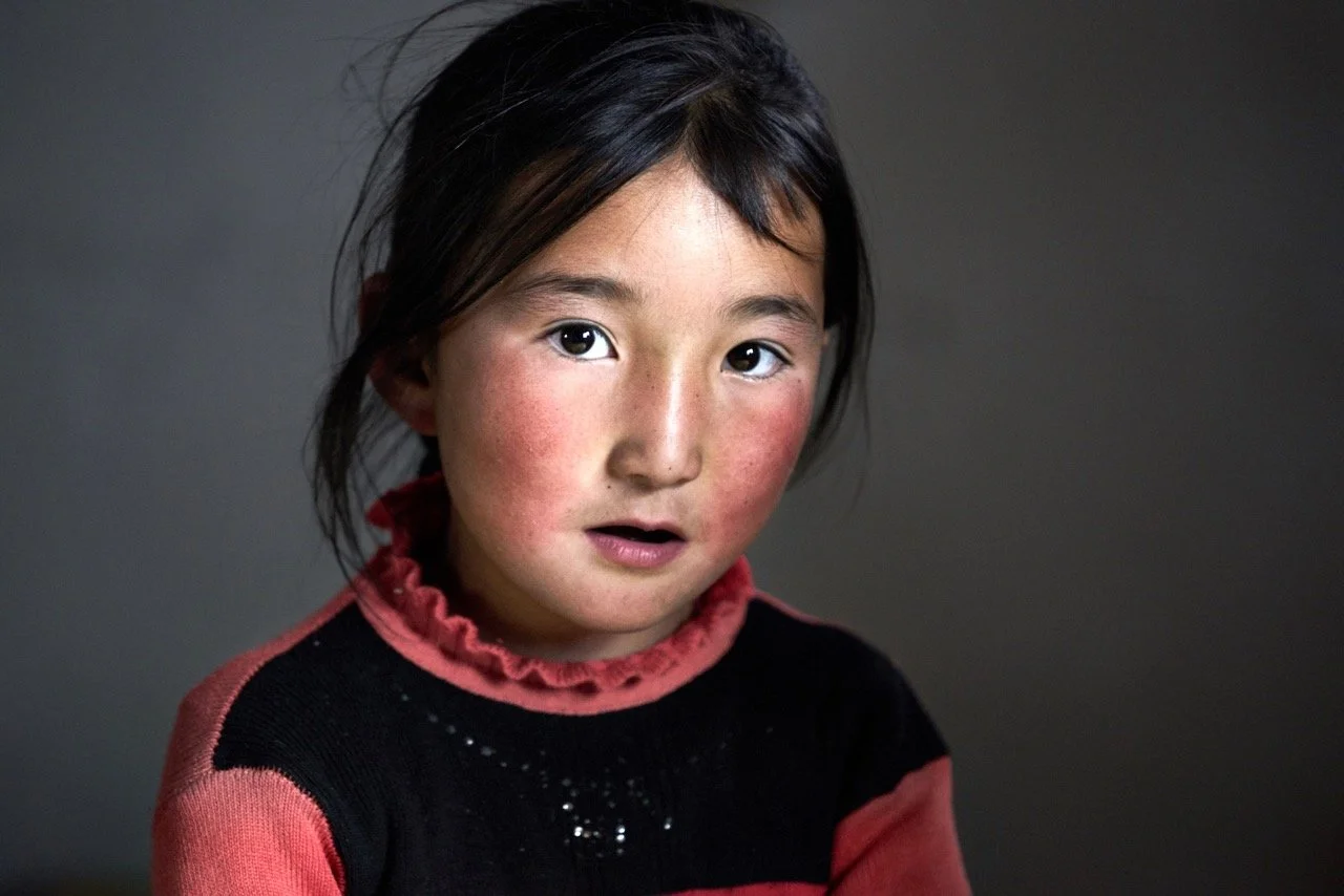 Young child with black hair and red cheeks, wearing a black and red sweater, looking at the camera.Film Mongolia. Filming in Mongolia. Producer in Mongolia. Film Production Service in Mongolia. Fixer Service Mongolia.
