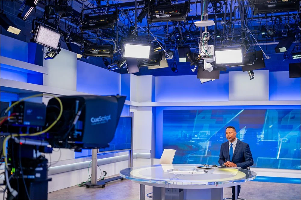 Wide view of broadcast news studio with anchor at desk, lighting grid, and camera equipment
