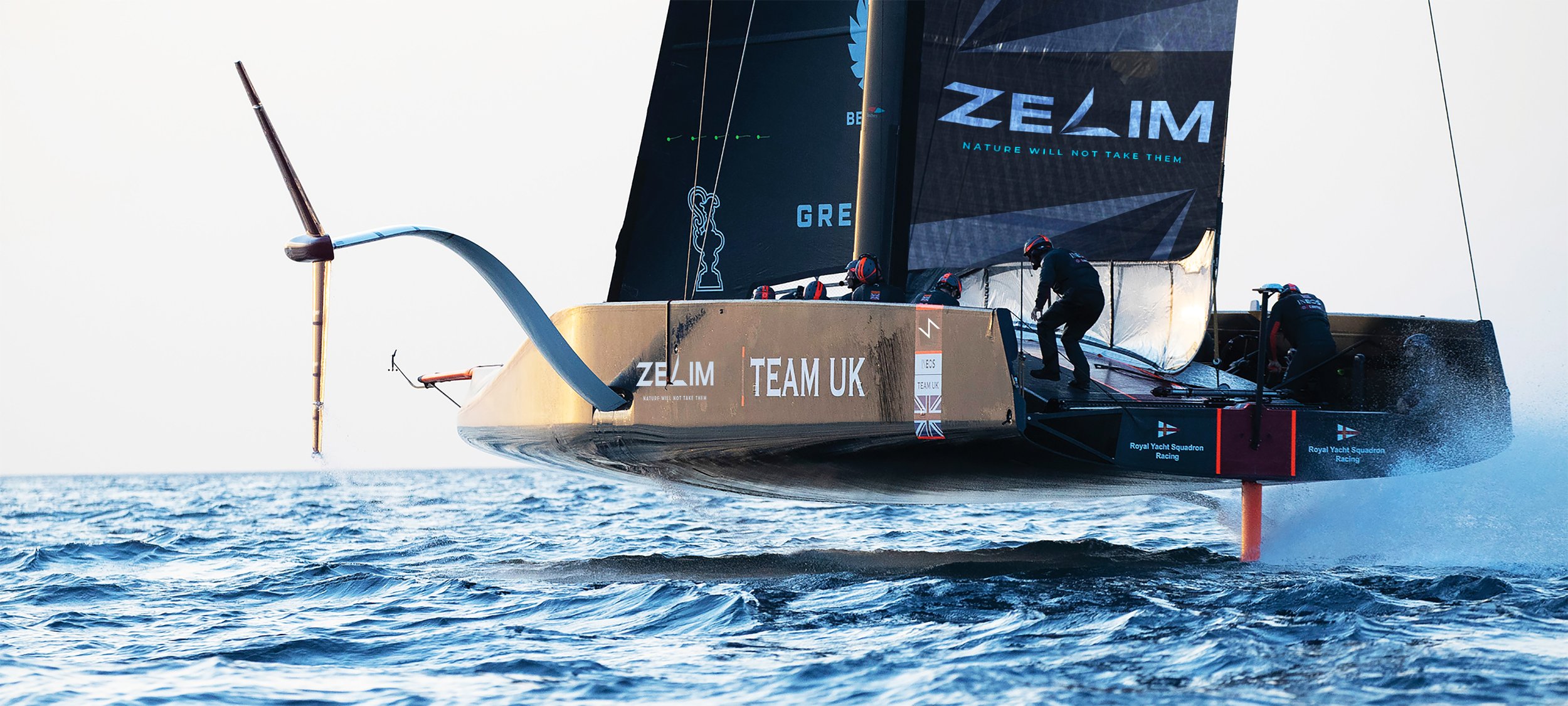 A racing sailboat with a crew of sailors on the water, black sails with logos, and the words 'Team UK' and 'ZELIM' visible. The boat is moving at high speed, creating waves and spray.