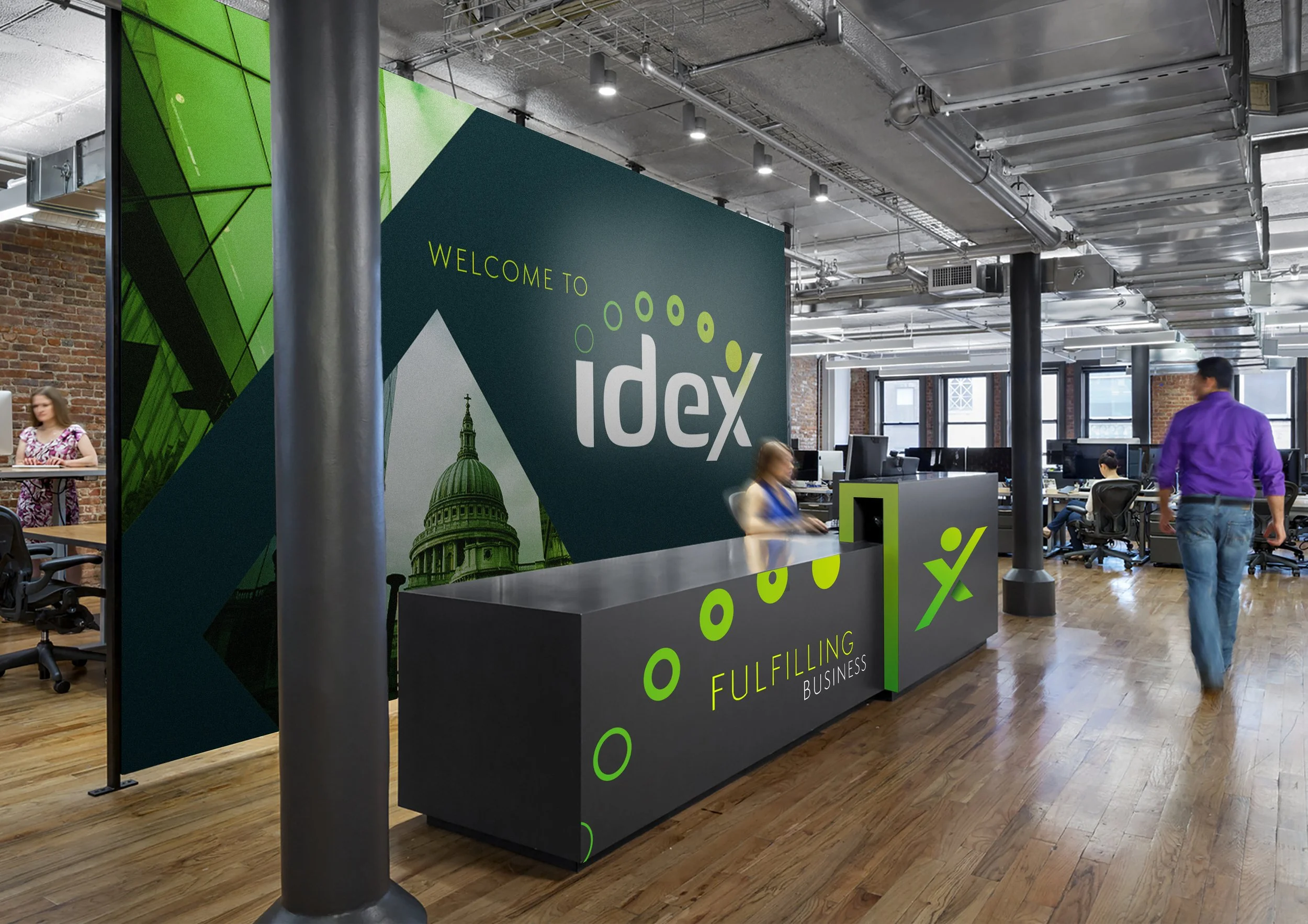 The image shows an office reception desk with a large welcome sign reading 'Welcome to IDex' and additional text 'Fulfilling Business'. The sign features a cityscape and green circular designs, set in a modern industrial office with exposed ductwork,