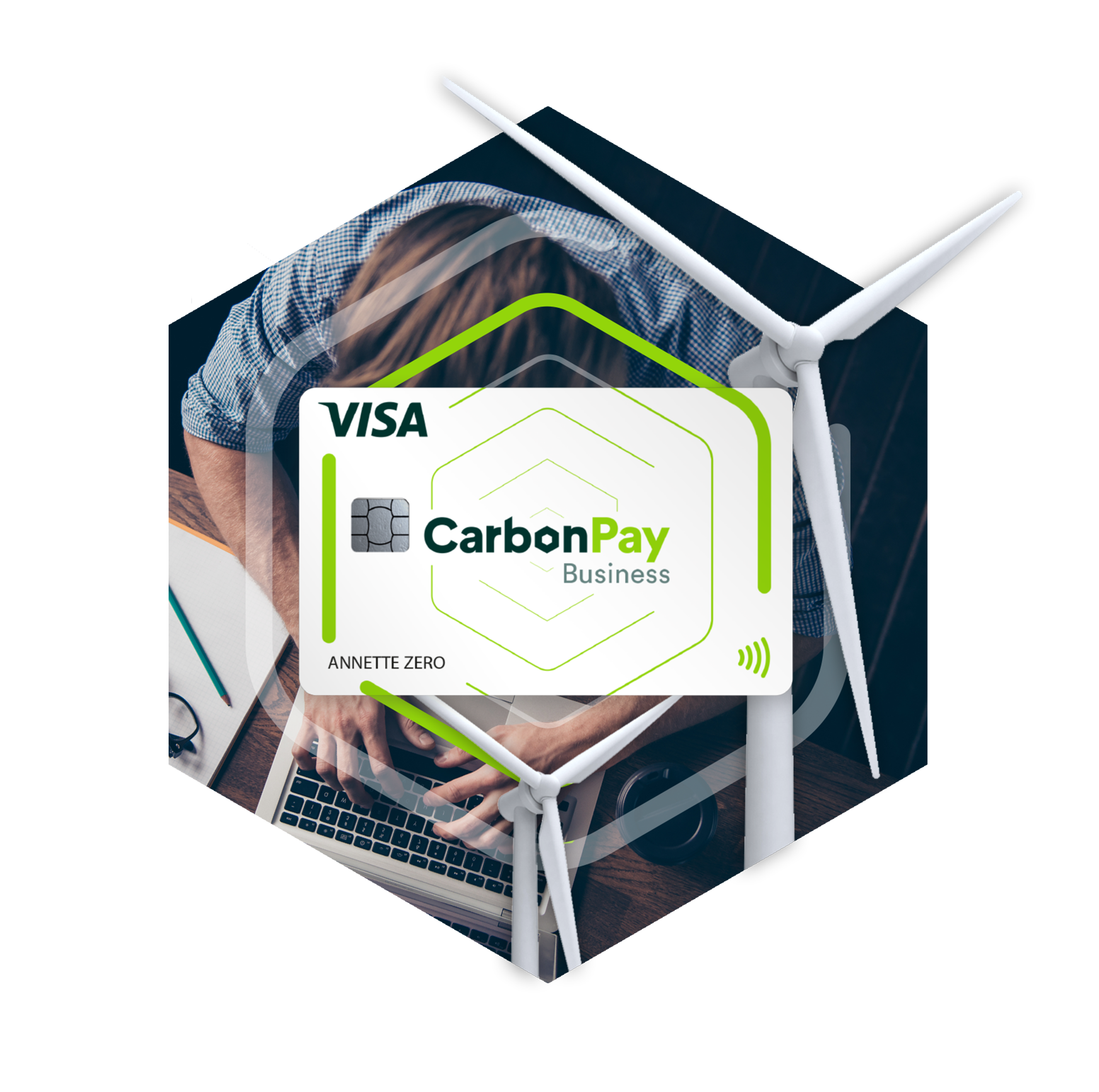 A person sitting at a desk, holding a smartphone displaying a Visa Carbon Pay card, with a laptop underneath and a white lamp nearby, in a modern office setting.