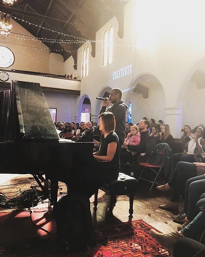 A woman plays piano and a man sings into a microphone on a stage in front of an audience in a church or auditorium with hanging string lights and stained glass windows.