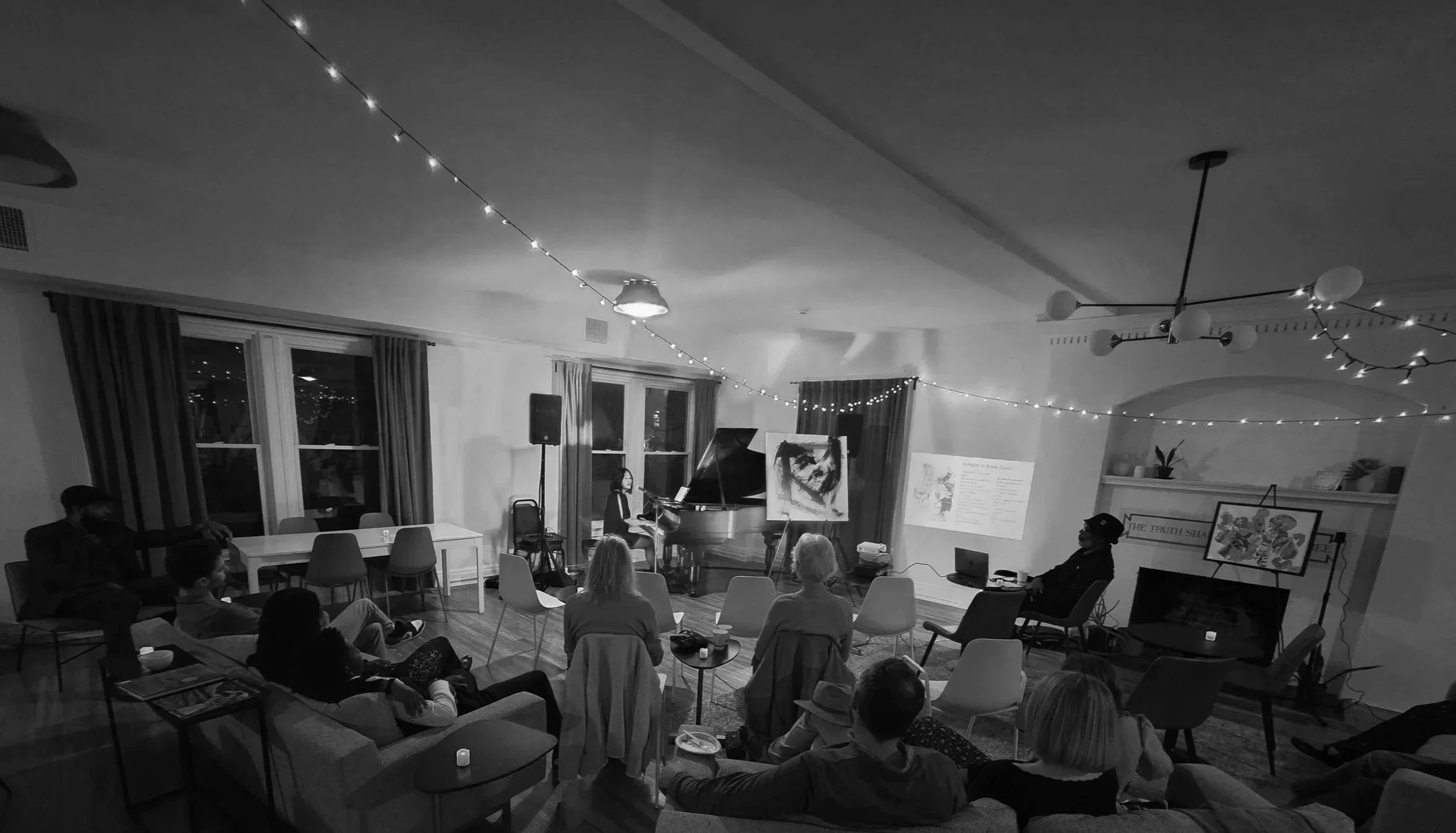 An indoor gathering with people sitting on chairs, watching a woman playing a grand piano, with a projection screen displaying text. Decor includes string lights and artwork on the walls.