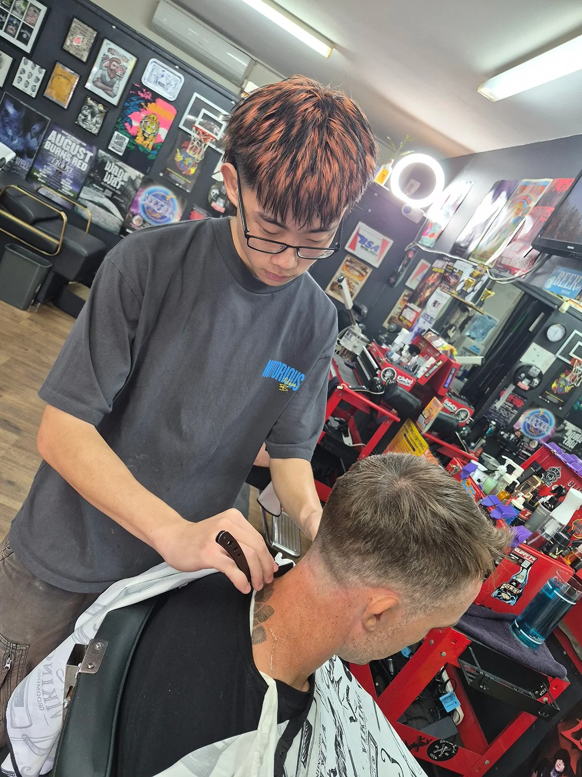 A person getting their hair cut at a barbershop by a barber wearing glasses and a black shirt, with barber tools and products visible on the counter and posters on the walls.