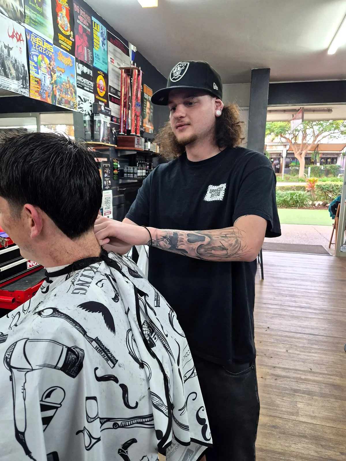 A barber cutting a male customer's hair inside a barbershop. The barber has long curly hair, tattoos on his arms, and is wearing a black t-shirt and a black cap. The customer is wearing a barber cape with black and white patterns. The background has posters on the wall and a view outside through open doors with trees and a sidewalk.