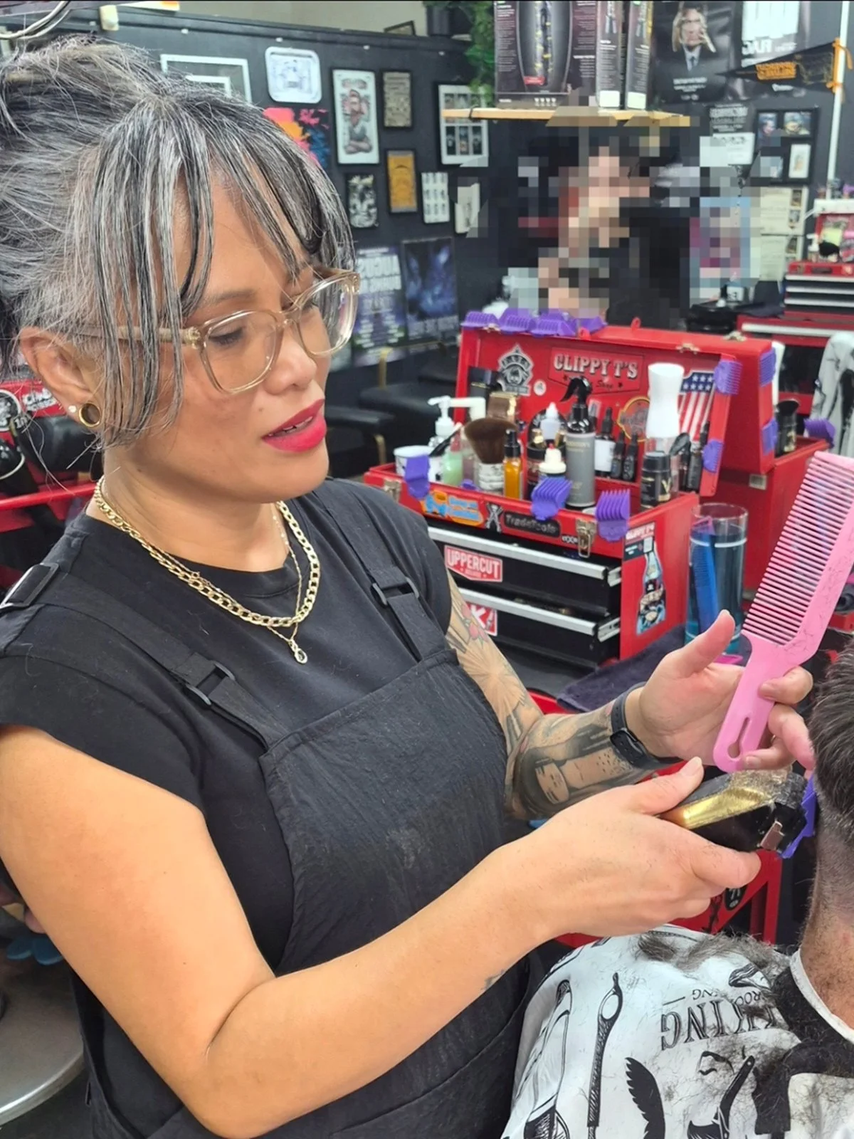 A woman with gray hair and glasses brushing a man's hair with a pink wide-tooth comb in a barber shop. The woman has tattoos on her arms, is wearing a black shirt and apron, and is holding a smartphone. The background shows framed pictures and a red toolbox filled with barber supplies.