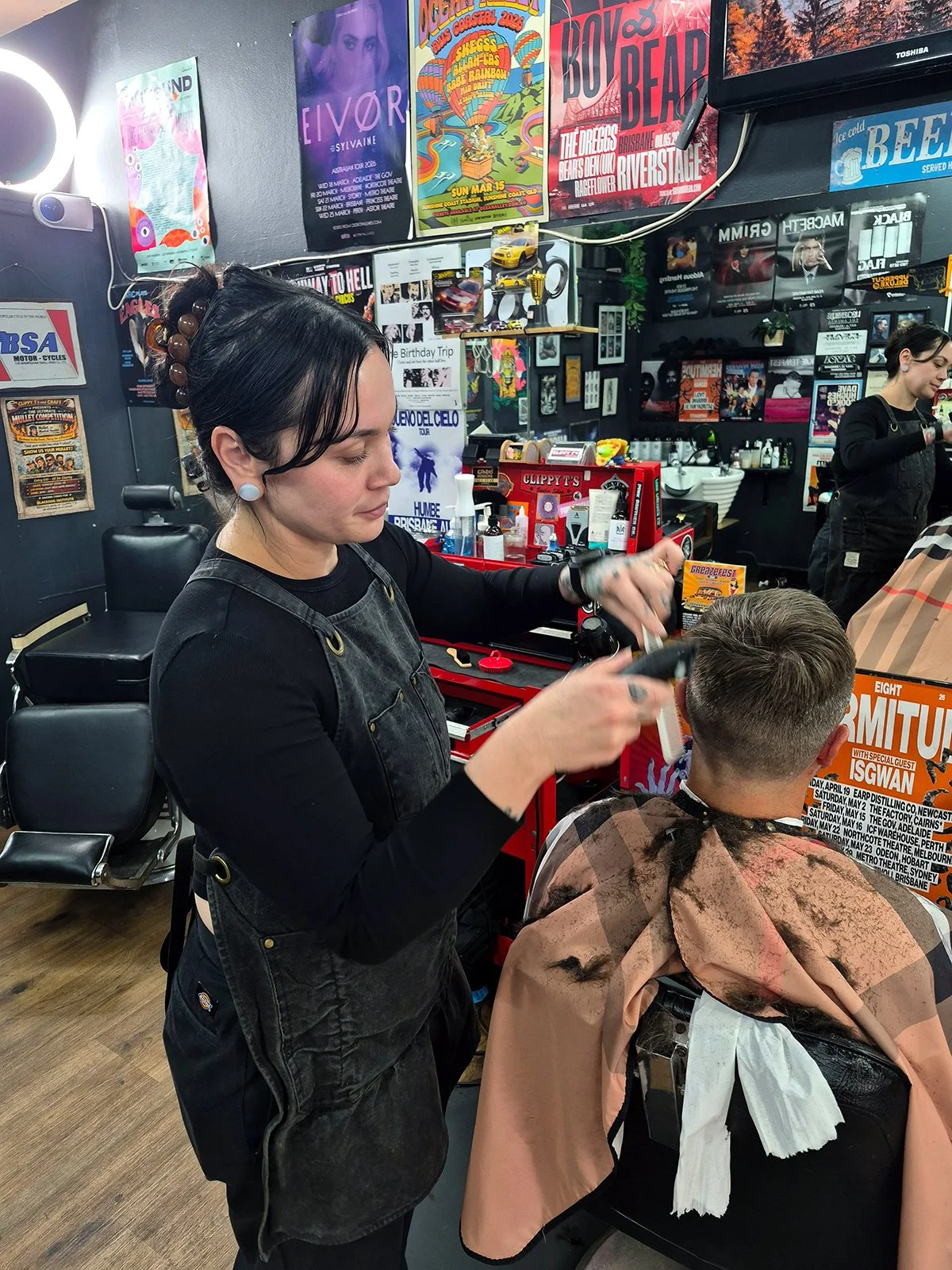 A hairstylist cutting a client's hair in a barbershop, with posters and artwork on the wall behind them.