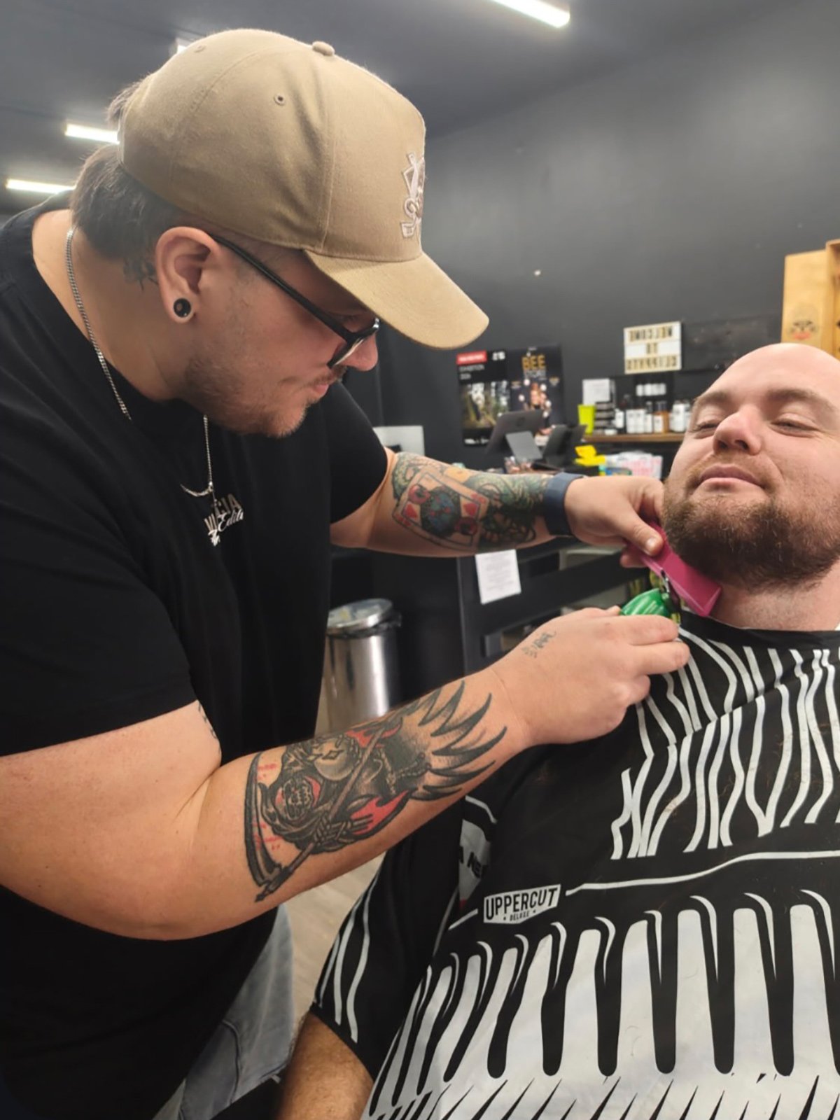 A barber wearing a beige cap, black shirt, and glasses is using an electric clipper to shave a man's neck. The man receiving the shave has a beard, is smiling, and is wearing a black and white striped barber cape. The background shows shelves with barber supplies and black walls.