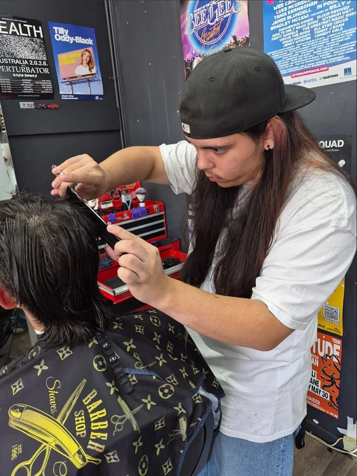 A barber with long hair, a black cap, and earrings is cutting a male customer's hair, who is seated, wearing a black cape with yellow patterns, in a barbershop. The background has posters and a red toolbox with grooming tools.