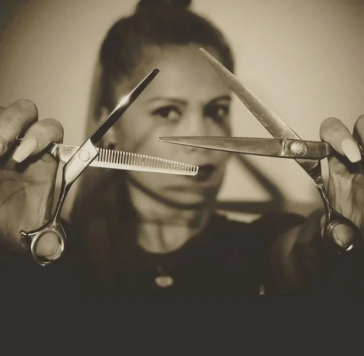 Black and white photo of a woman holding a pair of scissors and a comb, with the scissors open and crossing each other in front of her face, framing her eyes.