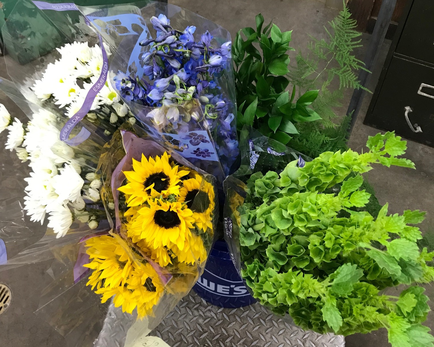 Small sunflowers, purple delphiniums, white pom pom daisies, green bells of Ireland, plumosa fern and Israeli ruscus.